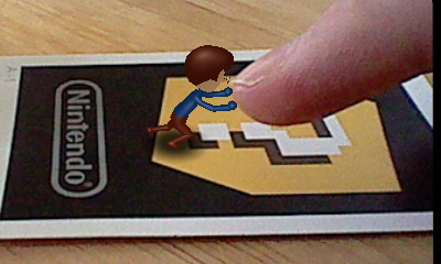 A picture of a miniature Nintendo Mii version of Tylor Lilley leaning against a giant finger, taken with a Nintendo 3DS and AR card