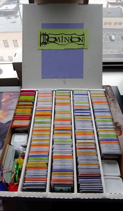 A picture of a beat-up cardboard BCW Super Monster storage box containing all Dominion cards released before Renaissance sleeved and organized within.